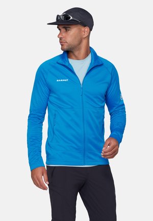 Mand iført blå fleecejakke med lynlås og "Mammut"-logo, sort kasket, solbriller på kasketten, lyseblå skjorte og mørke bukser, ser mod venstre.