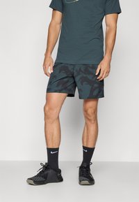 Herre sportstøj bestående af en mørkegrøn t-shirt, sorte mønstrede shorts og sorte sneakers med grå detaljer samt ankelsokker.