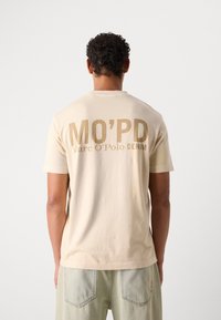 Beige cotton t-shirt with short sleeves, featuring bold brown text "MO’PD Marc O’Polo DENIM" on the back. Simple and relaxed design.