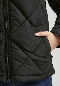 Svart quiltad jacka med dragkedja, diagonalt sömnadsmönster och sidofickor. Slät tyg med en något glänsande yta.