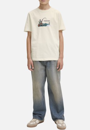 T-shirt col rond en beige clair avec un imprimé graphique. Coupe ample, associé à un jean large délavé bleu et des baskets beiges.