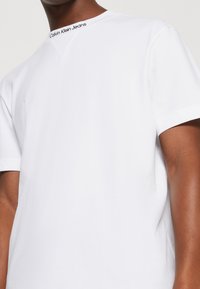Camiseta blanca de manga corta hecha de algodón con cuello redondo. Presenta el logo "Calvin Klein Jeans" en negro en el cuello. Textura suave.