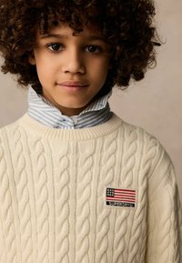 Cremfarbener Pullover mit Kabelstrickmuster und geripptem Kragen, versehen mit einem mehrfarbigen Patch, das eine amerikanische Flagge und das Markenlogo auf der linken Brust zeigt.