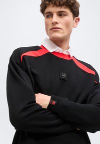 Νεαρός με μπούκλες, που φοράει μαύρο φούτερ με κόκκινες ρίγες και λευκό γιακά, με σταυρωμένα τα χέρια, κοιτάζει προς τα δεξιά του.