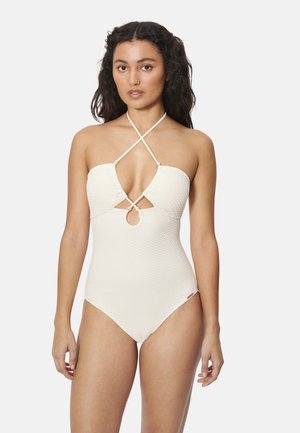 Mujer con cabello largo y rizado, vistiendo un traje de baño blanco de una pieza con textura, tirantes tipo halter cruzados y un detalle de recorte en el pecho, de pie sobre un fondo blanco.
