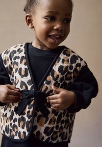 Chaleco de estampado de leopardo con ribete negro, que cuenta con un cierre de lazo, llevado sobre una sudadera negra. El material parece suave con una textura acolchada.