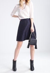 Námořnicky modrá áčková sukně s jemným geometrickým vzorem, doplněná světlobéžovou blůzou, černou kabelkou s třásněmi a kotníkovými botkami.
