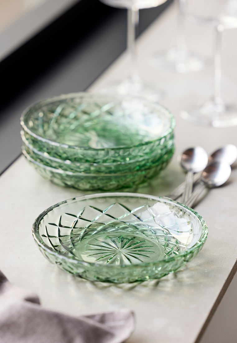 Zielone, szklane miski z misternymi wzorami diamentowymi i gwiazdkowymi, ułożone jedna na drugiej, na jasnej powierzchni z srebrnymi łyżkami obok.