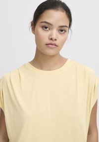 Camicia gialla a maniche corte con vestibilità rilassata e tessuto morbido, caratterizzata da accenti plissettati sulle spalle e un collo arrotondato.