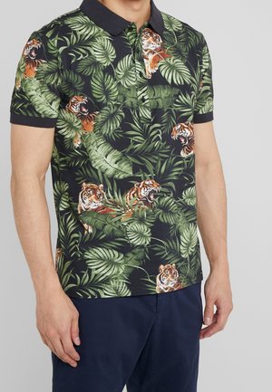 Mann trägt ein schwarzes Poloshirt mit Kragen, bedruckt mit grünen tropischen Blättern und einem brüllenden Tiger, kombiniert mit dunkelblauen Hosen.