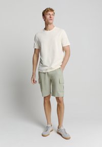 Lichtbeige katoenen t-shirt en lichtgroene cargoshorts met zakken, gecombineerd met grijze sneakers, staande op een neutrale achtergrond.