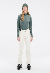 Pantalons de ski blancs légers avec poches zippées, associés à un haut à manches longues vert texturé avec des rayures verticales et un bonnet en tricot assorti.