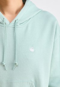 Sweat à capuche vert menthe en tissu doux, avec une poche avant et un petit logo blanc sur la poitrine. Cordons ajustables au niveau de la capuche.