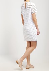 White patterned dress with a round neckline and short sleeves, featuring a small back keyhole detail, paired with beige flat shoes.