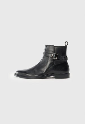 Botas de cuero negro hasta el tobillo con puntera redondeada, detalle de hebilla lateral y una pestaña en el talón. Suela plana con un acabado sutilmente brillante.