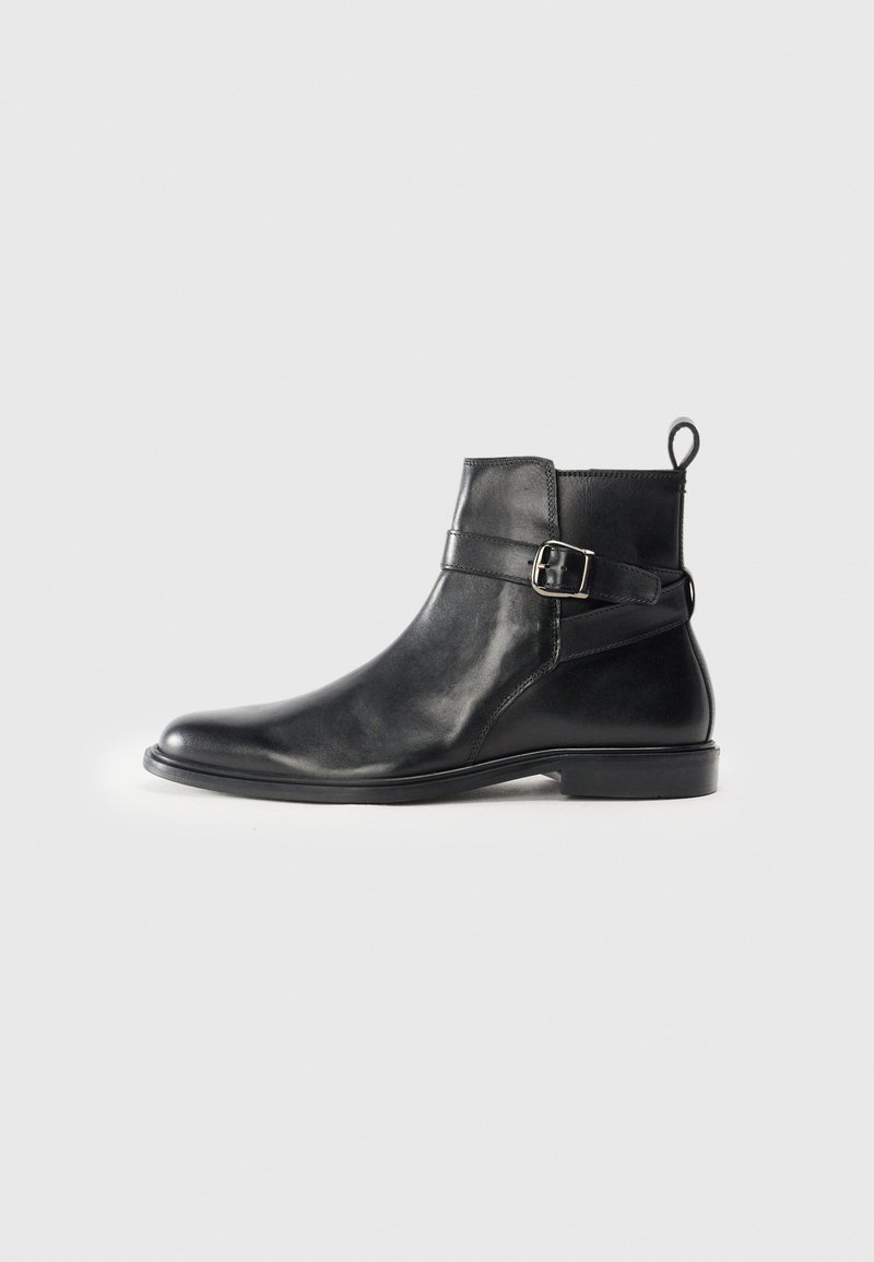 Botas de cuero negro hasta el tobillo con puntera redondeada, detalle de hebilla lateral y una pestaña en el talón. Suela plana con un acabado sutilmente brillante.