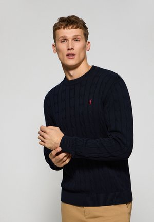 Marineblauer Strickpullover mit Zopfmuster, gerippten Bündchen und Saum, kleinem roten Logo auf der Brust, Rundhalsausschnitt, kombiniert mit hellbeigen Hosen.