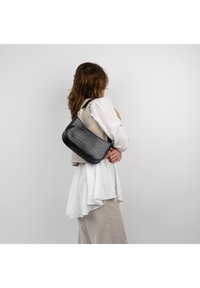 Schultertasche im schwarzen Krokodilmuster mit goldenen Hardware-Details, die eine weiche Textur und eine strukturierte Form aufweist, getragen über einem leichten, geschichteten Outfit.