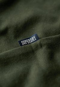 Tessuto verde oliva con una texture liscia e un primo piano di un'etichetta blu con la scritta "SUPERDRY" in lettere maiuscole bianche.