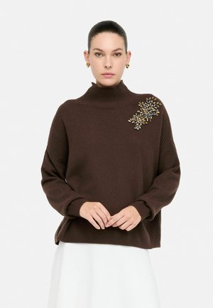 Maglione marrone lavorato a maglia con colletto alto a coste e un'applicazione floreale blu e oro sulla spalla sinistra. Texture morbida, vestibilità comoda.