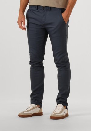Marineblauwe slim-fit broek van een glad materiaal met zijzakken, gecombineerd met beige en witte sneakers met textuurdetails.