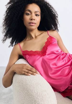 Camisole en satin rose avec une dentelle rouge le long du décolleté. Le modèle pose sur un canapé crème texturé, mettant en valeur la finition lisse du vêtement.