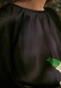 Blouse noire en voile avec un col froncé, ornée d'un motif de fleur blanche et de feuilles vertes sur le côté inférieur droit. Texture lisse.