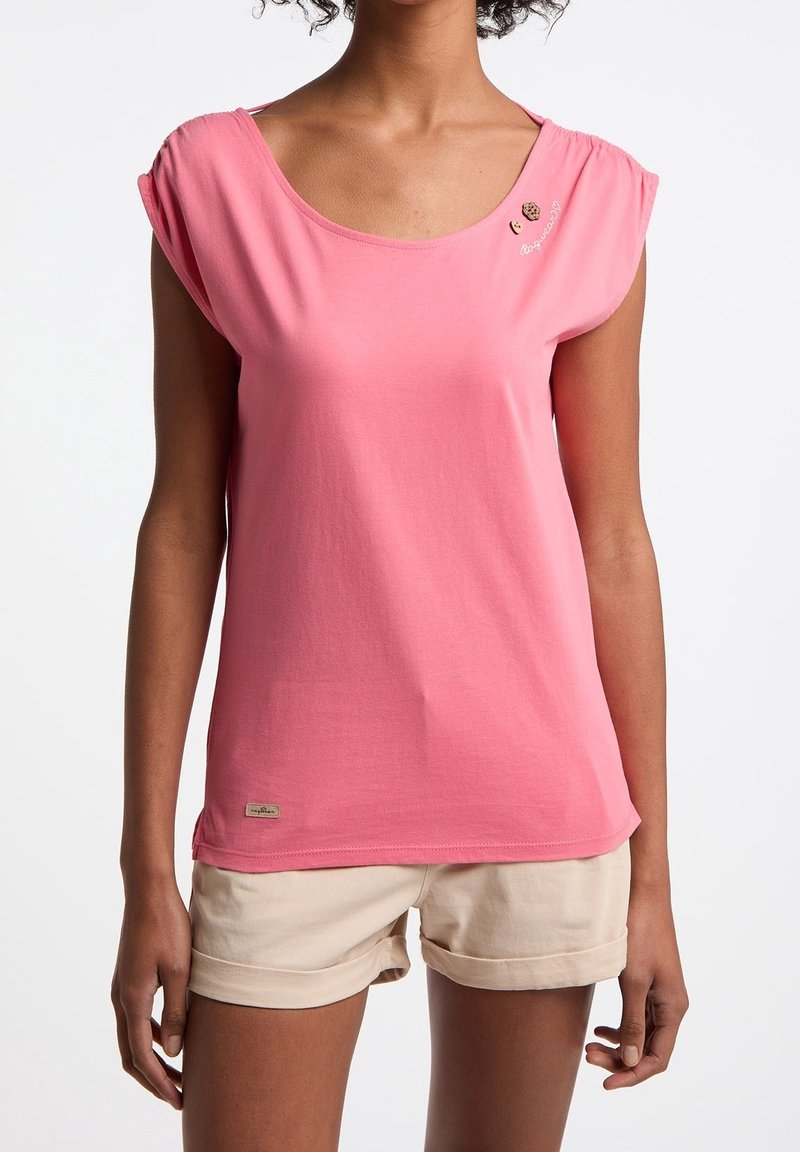 Rosa T-Shirt mit kurzen Ärmeln und Rundhalsausschnitt, aus weichem Baumwollstoff, mit dezentem Stickerei an der Schulter. Kombiniert mit beigen, hochgekrempelten Shorts.