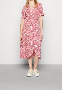 Robe midi rouge à motif cachemire avec manches courtes bouffantes, un décolleté en V profond et un design portefeuille, associée à des baskets blanches.