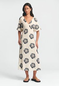 Blommig midiklänning i krämfärgad tyg, med V-ringning, korta klockärmar och marinblå blommönster överallt. Modellen bär svarta sandaler.