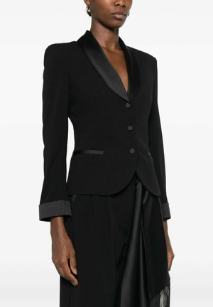 Donna che indossa un blazer nero aderente con revers e polsini in raso, chiusura con bottoni frontale, e pantaloni neri abbinati con dettaglio in raso drappeggiato.