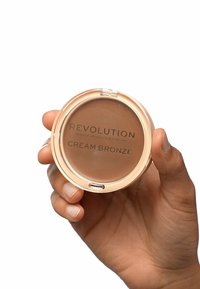 Compact rond et transparent contenant un produit crème bronzé. L'étiquette indique "REVOLUTION CREAM BRONZE." Une main tient le compact, mettant en avant la texture lisse.