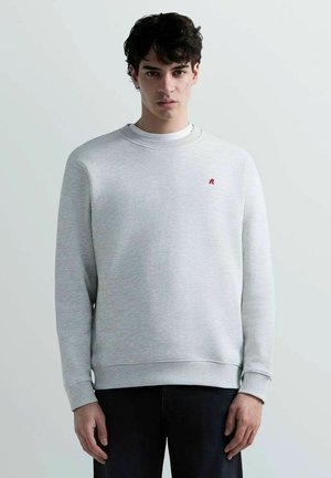 Giovane uomo con capelli scuri che indossa una felpa girocollo grigio chiaro con un piccolo logo rosso sul petto, in piedi contro uno sfondo chiaro e uniforme.