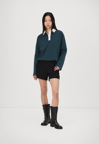 Tmavomodrý polo tričko s dlhými rukávmi a bielym golierom, v kombinácii s čiernymi šortkami a čiernymi ponožkovými čižmami. Stojí na bielom pozadí.