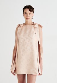 Frau, die ein ärmelloses, blassrosa Minikleid mit floralem Jacquardmuster und langen Schleifen an den Schultern trägt, vor einem schlichten weißen Hintergrund.