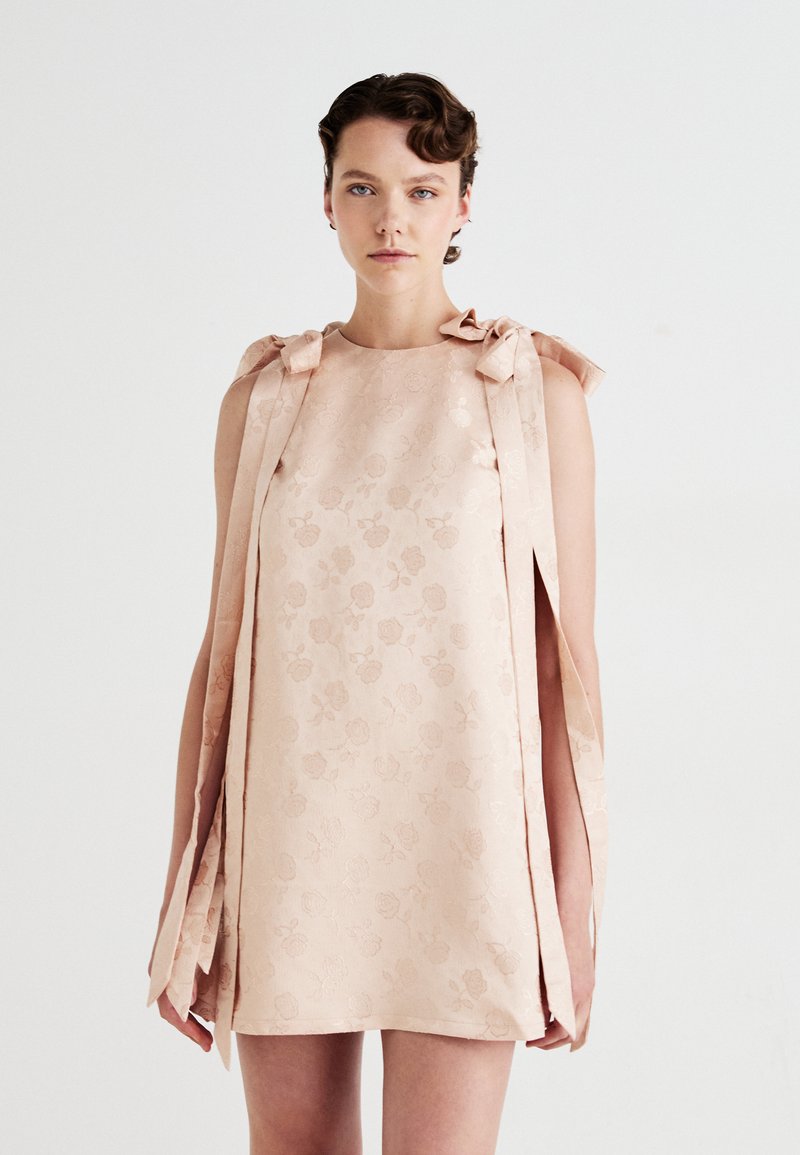 Frau, die ein ärmelloses, blassrosa Minikleid mit floralem Jacquardmuster und langen Schleifen an den Schultern trägt, vor einem schlichten weißen Hintergrund.