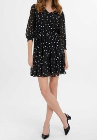 Robe noire à pois blancs avec col en V, manches trois-quarts, taille cintrée et jupe à volants en tissu léger. Talons noirs assortis.