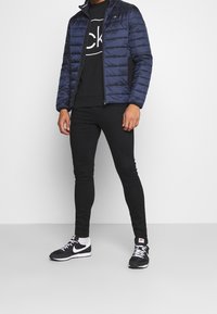 Námorně modrá prošívaná bunda s vysokým límcem, černé grafické tričko, úzké černé kalhoty a černé tenisky Nike s bílými akcenty.