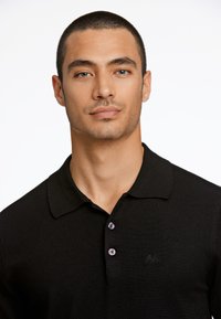 Camisa polo de algodón negra con collar clásico, que presenta dos botones morados y un logotipo bordado en el pecho. Textura suave.