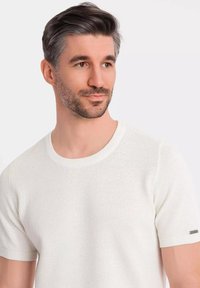 Suéter corto de manga corta texturizado en blanco, con un escote redondo y un sutil acento metálico en la manga. Material de tejido ligero.