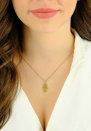 Collier en pendentif en or présentant un symbole de main avec des motifs élaborés, sur une fine chaîne en or, porté sur une blouse blanche.