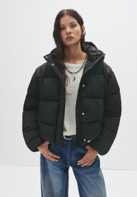 Jonge vrouw in een zwarte gewatteerde jas, witte blouse, blauwe spijkerbroek en gelaagde zilveren kettingen, staande tegen een eenvoudige witte achtergrond.