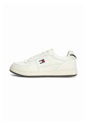 Baskets blanches basses avec un design à lacets, logo Tommy Jeans noir et rouge sur le côté, et languette de talon rayée noire.
