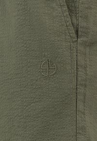 Primo piano di un tessuto verde oliva testurizzato con un sottile logo circolare ricamato vicino a una cucitura verticale.