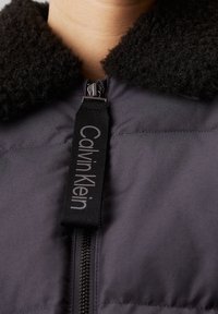 Zbliżenie na czarną kurtkę zapinaną na zamek z puszystym kołnierzem i czarnym uchwytem do zamka błyskawicznego z logo Calvin Klein.