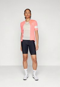 Roze fietsjersey met korte mouwen en witte accenten over een doorzichtige binnenlaag, gecombineerd met zwarte fietsbroek en witte sportkousen.