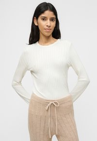 Γυναίκα με μακριά σκούρα μαλλιά, φορώντας λευκό ριμπ μπλουζάκι με μακριά μανίκια και μπεζ πλεκτό παντελόνι με δέσιμο μπροστά, στέκεται μπροστά σε απλό ανοιχτόχρωμο φόντο.