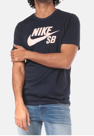 Homme portant un t-shirt Nike SB bleu marine et un jean bleu clair, avec un bras croisé tenant l'autre bras.