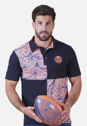 Homme portant un polo bleu marine avec des panneaux à motifs orange et tenant un ballon de rugby assorti à hauteur de taille.