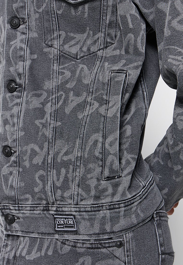 Chaqueta de mezclilla en gris oscuro con un patrón texturizado, cierre frontal a botones y bolsillo tipo parche con etiqueta de marca.